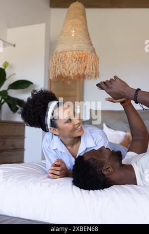 Young diverse couple relax on bed looking at a smartphone. cozy bedroom with wooden furniture and large woven lamp in background, unaltered Stock Photo