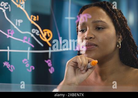 Biracial young woman writing on transparent board with orange marker in an business office. Graphs and charts in background, creating focused and prof Stock Photo