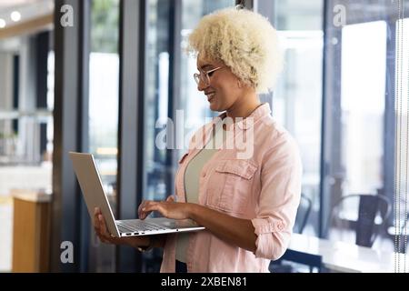 Young biracial woman using laptop, smiling while working in business office . Large windows and modern furniture creating bright and open workspace, u Stock Photo