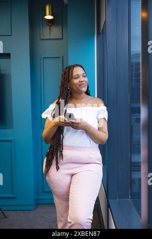 Young biracial woman smiling, holding smartphone by window in modern business office. Teal background and large windows creating bright and open atmos Stock Photo
