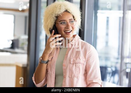 Biracial woman with curly hair, glasses, in casual wear, talking on smartphone. with glass walls and modern furniture in background, unaltered Stock Photo