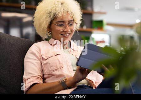 Young biracial woman with curly hair using tablet, smiling in a modern business office. Plants and shelves in background, creating productive atmosphe Stock Photo