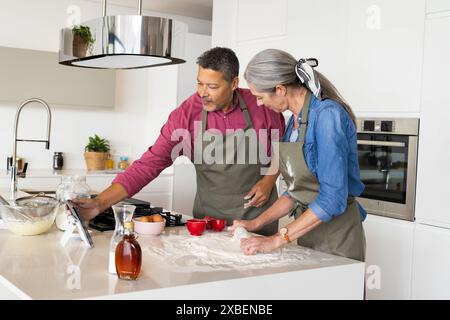 A biracial mature couple cooks together in a modern kitchen, at home. They are preparing dough on a white countertop with various ingredients around t Stock Photo