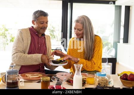A biracial mature couple is enjoying breakfast together at home, at home. They are in bright, modern kitchen with various breakfast items on table, un Stock Photo