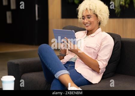 Young biracial woman, team member, using tablet and smiling in business office lounge area. Plants and coffee cup nearby, creating relaxed and product Stock Photo