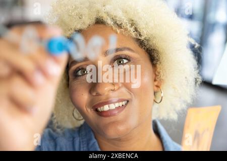 Biracial young woman writes on glass board, at camera smiling in business office. undefined environment with blurred colleagues in background, fosteri Stock Photo