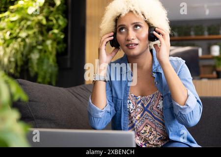 A young biracial woman with curly blonde hair wears headphones on a couch in a modern business offic Stock Photo