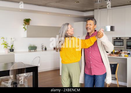 A biracial mature couple is dancing together in modern kitchen, at home. The bright and spacious kitchen features sleek white cabinets and contemporar Stock Photo