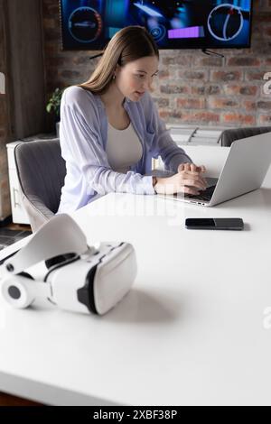 Young Caucasian woman working on laptop in modern office with brick background. Tech gadgets creating high-tech workspace, enhancing productivity and Stock Photo