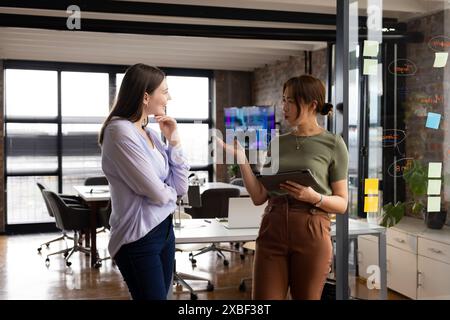 Asian businesswoman, discussing project with young caucasian woman. modern office with glass walls and sticky notes on board in background, unaltered Stock Photo