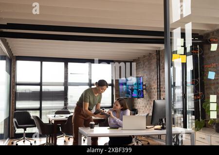 Asian businesswoman showing something on tablet to young Caucasian woman in modern office. Brick walls, large windows, and screen displaying code crea Stock Photo