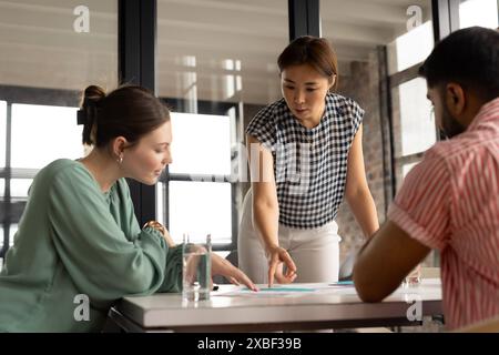 Young Caucasian woman and Asian man discussing project with Asian businesswoman. Modern office with large windows and natural light enhances collabora Stock Photo