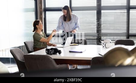 Asian businesswoman shaking hands with young Caucasian woman in modern office. Large windows with blinds and conference table with office supplies cre Stock Photo