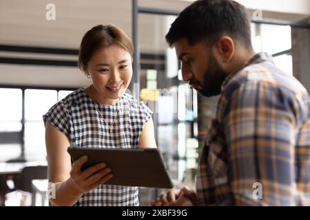 Asian businesswoman showing tablet to Asian male colleague in office. Modern workspace with large windows and natural light filling room, unaltered Stock Photo