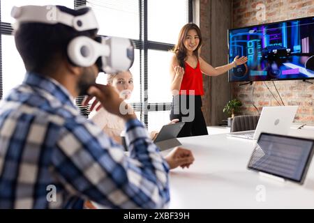 Asian businesswoman presenting to team with VR headset and tablet. Modern office with large windows and brick walls, team members paying close attenti Stock Photo