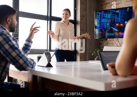 Asian businesswoman presenting to diverse team in a modern office. Large screen displaying a vibrant, futuristic interface with colorful graphics, una Stock Photo