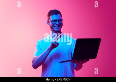 Smiling young man in glasses and white t-shirt holding laptop and having online video call with friends against pink background in neon light Stock Photo