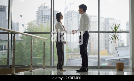 Two business partners colleagues people shaking hands in office Caucasian woman and man handshaking greeting talking in lobby corridor cooperation Stock Photo