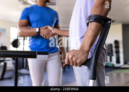 Biracial man with crutches shaking hands with male physical therapist. They are in rehabilitation center with exercise equipment and weights in backgr Stock Photo