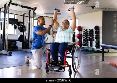 Quadriplegic biracial woman in wheelchair exercising with dumbbells, assisted by trainer they are in rehabilitation gym with various exercise equipmen Stock Photo