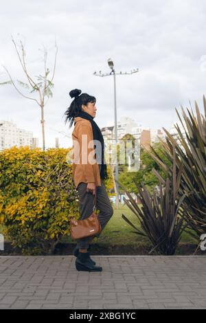 side view of a Elegance Latin Businesswoman walking down a sidewalk, holding a brown leather wallet. She has her hair in a bun. The scene is set in a Stock Photo