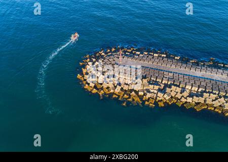 aerial zenithal view of a fishing boat leaving a harbor protected by breakwaters, drone image Stock Photo