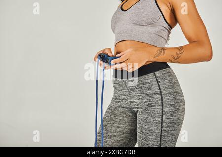 African American sportswoman in activewear energetically holds a jump rope in a studio with a grey background. Stock Photo