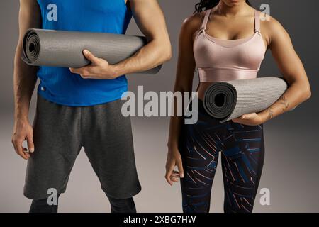 A young multicultural and fit couple in active wear holding a yoga mat in a studio setting against a grey background. Stock Photo