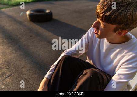 Young Man Sitting on Sidewalk With Tire in Background at Sunset Stock Photo