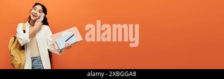 Young indian woman holding laptop, talking on phone against orange wall. Stock Photo