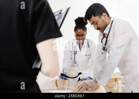 Two medical students in white coats practice CPR on a mannequin while a third student observes. Stock Photo