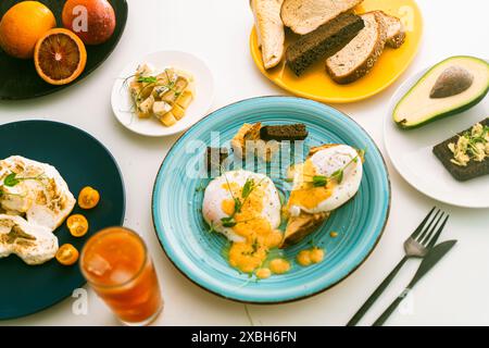 Eggs Benedict with hollandaise sauce and peas on a turquoise plate and avocado with rye toast, cheese, blood oranges and orange juice on a white table Stock Photo