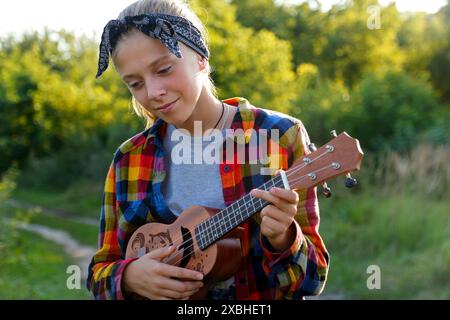 Portrait of a young girl playing ukulele outside, enjoying music. Girl playing guitar outdoors, ukulele in hand, portrait. Materials for music lessons Stock Photo