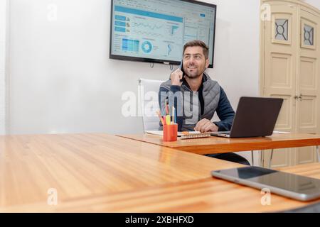 Middle-aged businessman talking on the phone while using the laptop on his desk, with his gaze to the side and smiling. Stock Photo