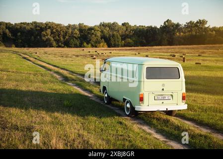 Green classic Volkswagen Transporter on field road driving away. Van on straight unpaved road amid hay stacks. Stock Photo