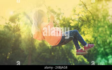 happy preschool girl swings high on summer day, blurred child figure in rays light, joyful Carefree childhood memories, boundless joy Stock Photo