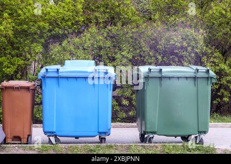 green, blue and brown trash can in front of green trees and bushes Stock Photo