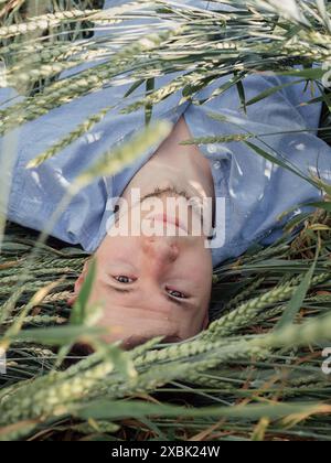Daydreaming: Vertical upside down portrait of a young man laying down on the green rye looking at camera. Stock Photo