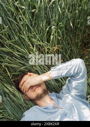 Daydreaming: Vertical straight from above close up shot of a young man in sky blue shirt laying on his back on the green rye, his hand covering the ey Stock Photo
