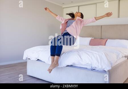 Happy biracial woman jumping on bed with arms outstretched, looking joyful Stock Photo