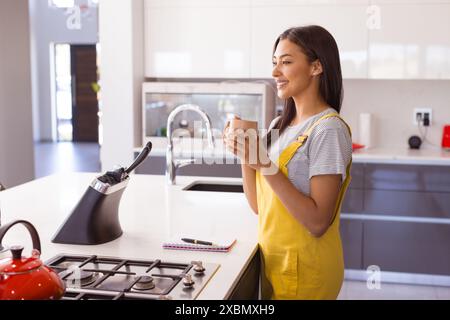 Happy biracial woman holding cup, standing in a modern kitchen, copy space Stock Photo