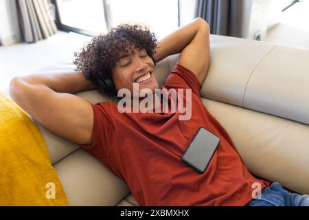 Happy biracial man relaxing on couch, smiling with a smartphone on his chest and headphones Stock Photo
