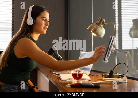 Smiling Caucasian woman wearing headphones using tablet at home, podcast recording Stock Photo