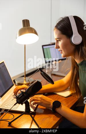 Smiling Caucasian woman with headphones recording podcast on laptop and microphone Stock Photo