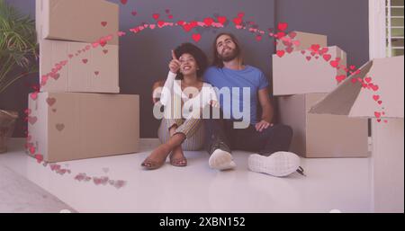Image of red hearts over happy diverse couple sitting amongst boxes after moving into house Stock Photo
