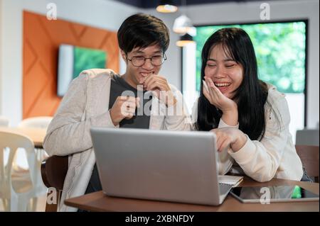 Two cheerful young Asian college students are laughing at something on a laptop computer, enjoying talking while working or studying together in a caf Stock Photo