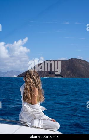 A woman sits on the bow of a boat, gazing out at a volcanic island in the distance. The water is a beautiful blue, and the sky is clear and sunny. Stock Photo