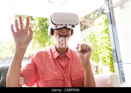 Senior caucasian woman using virtual reality headset, sitting on white armchair Stock Photo
