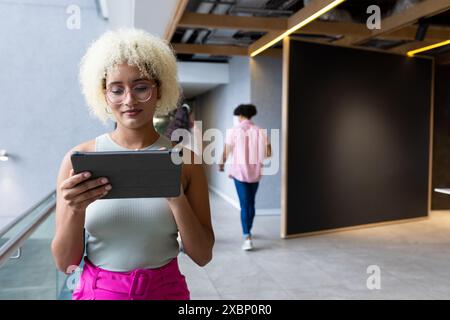 A biracial woman using tablet while walking in modern office hallway, business, copy space Stock Photo
