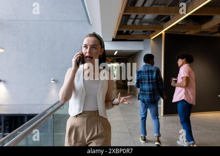 Caucasian woman talking on smartphone, walking in office hallway, business Stock Photo